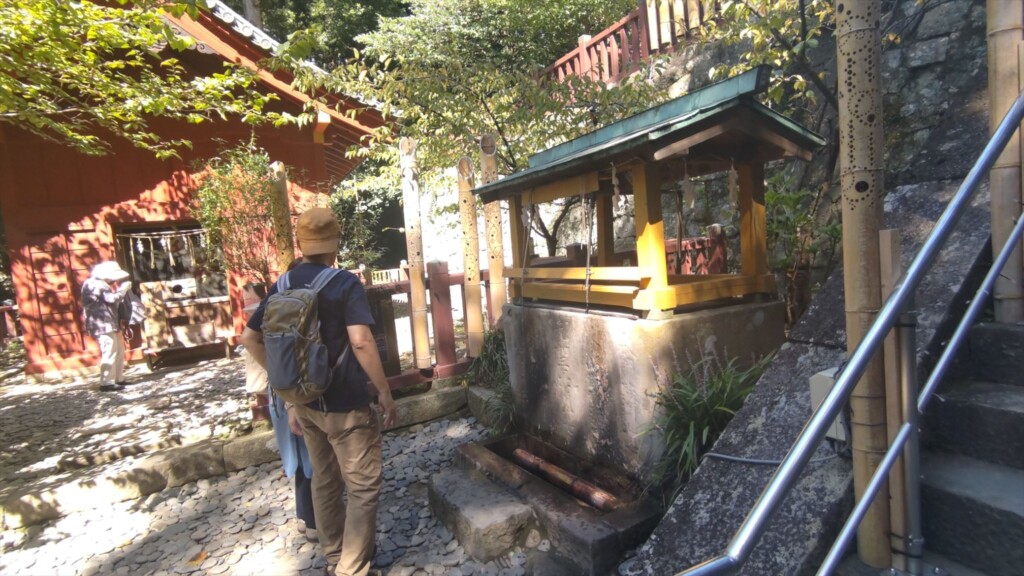 Tourist washing hands at the temizuya water basin at Kunozan Toshogu Shrine, with the historic Jingyu horse stable visible in the background.