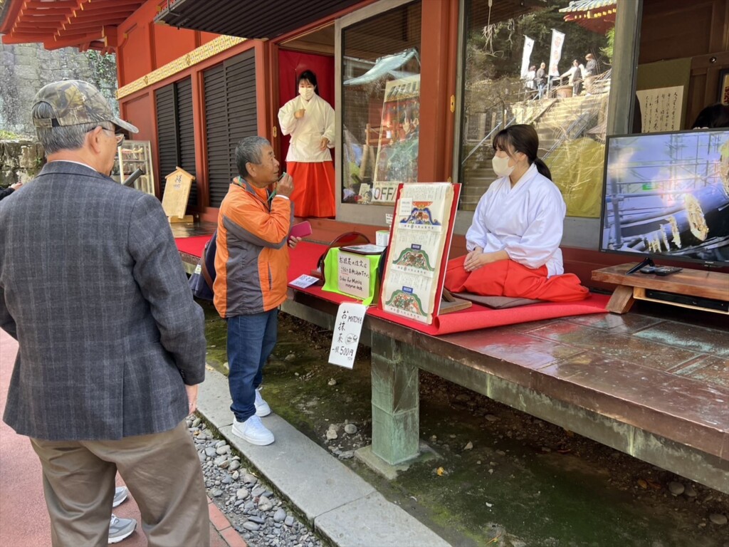A shrine maiden dressed in a traditional white kimono and red hakama is tolking with a tour guide.